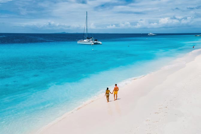 Paar spaziert am wei&szlig;en Sandstrand; t&uuml;rkisblaues Meer mit sanften Wellen, im Hintergrund ankern Segel- und Motorboote.