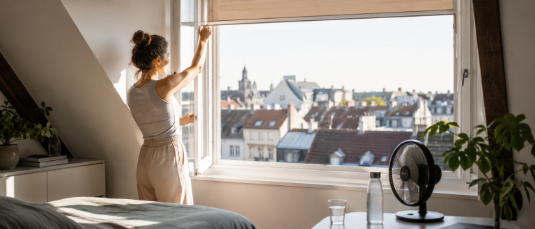 Person öffnet in einer Dachgeschosswohnung am Morgen das Fenster, daneben stehen Ventilator, Wasserflasche und Glas gegen die Sommerhitze.
